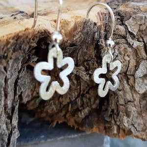 K&ouml;nnte beinhalten: Ein Paar silberne Blumenohrringe. Jeder Ohrring hat ein Blumendesign mit f&uuml;nf Bl&uuml;tenbl&auml;ttern und einer kleinen Kugel an der Spitze. Die Ohrringe werden auf einem St&uuml;ck Holz mit rauer Textur pr&auml;sentiert.