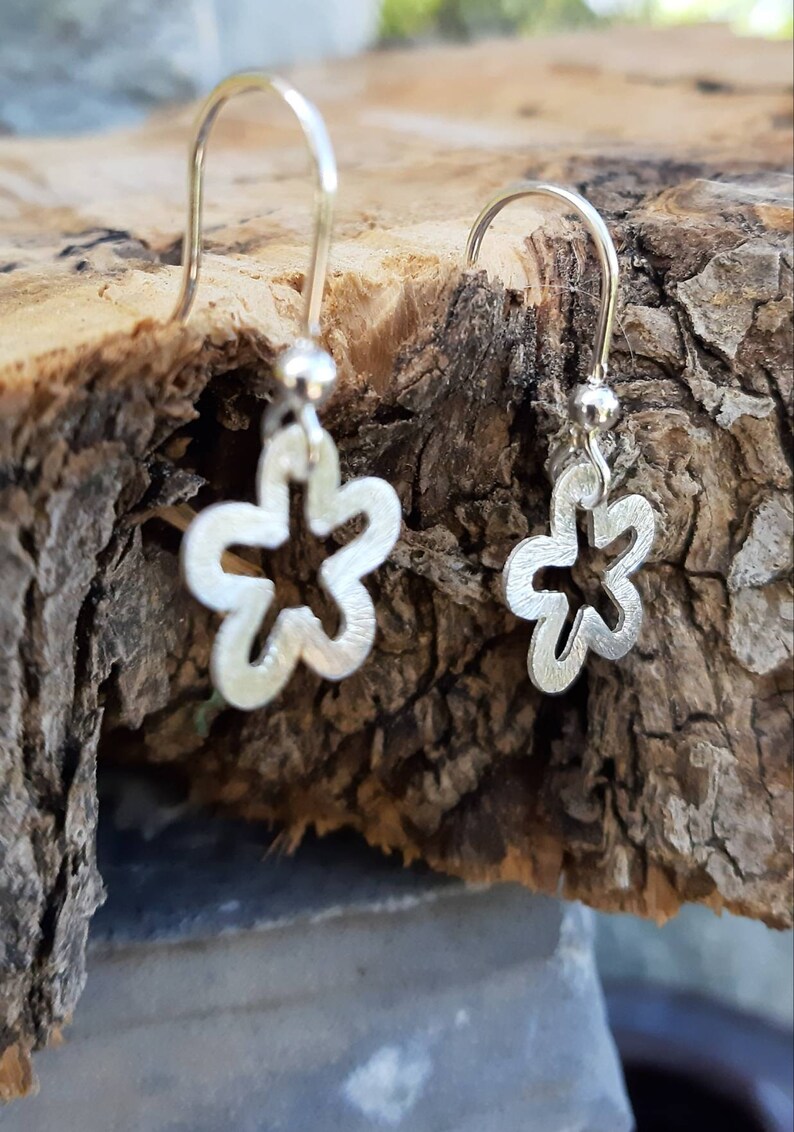 K&ouml;nnte beinhalten: Ein Paar silberne Blumenohrringe. Jeder Ohrring hat ein Blumendesign mit f&uuml;nf Bl&uuml;tenbl&auml;ttern und einer kleinen Kugel an der Spitze. Die Ohrringe werden auf einem St&uuml;ck Holz mit rauer Textur pr&auml;sentiert.