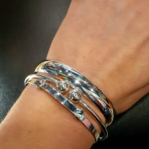 May include: Three silver bracelets stacked on a wrist. The top bracelet is a thick, rounded band. The middle bracelet is a thinner band with two small silver balls. The bottom bracelet is a thin, flat band.