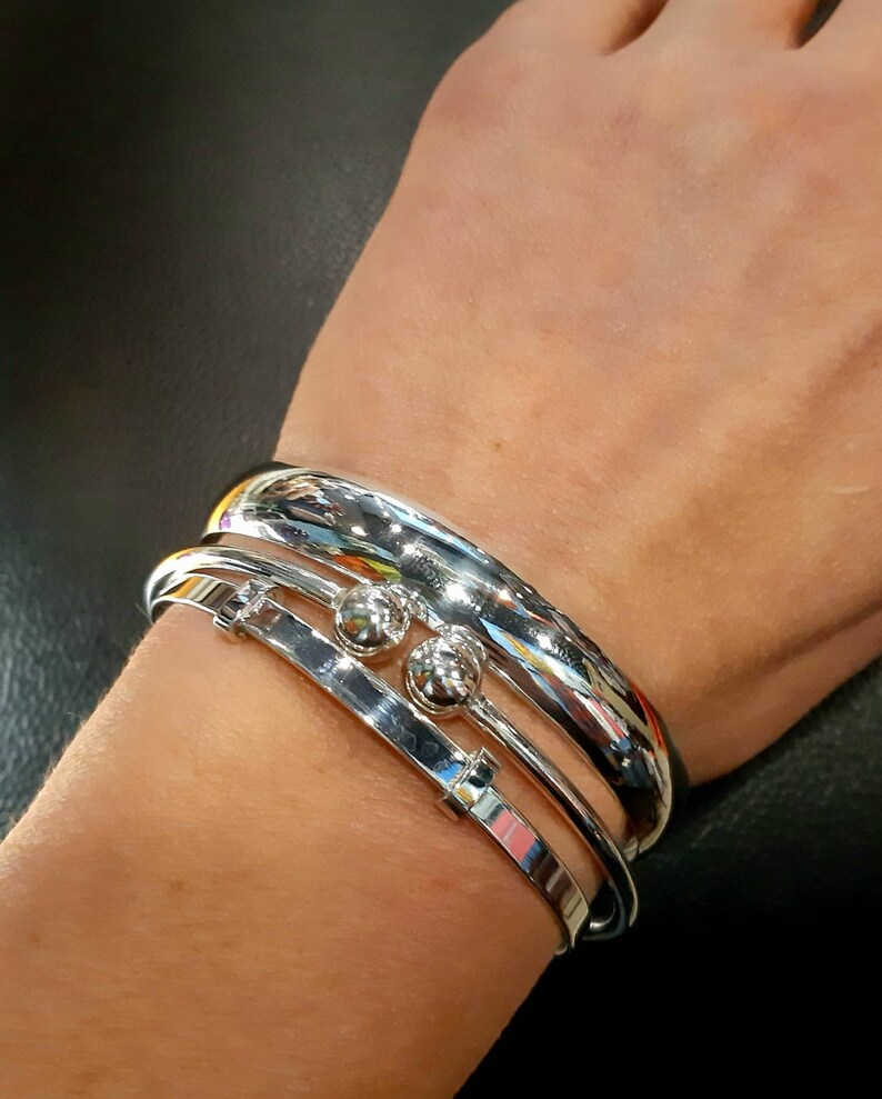 May include: Three silver bracelets stacked on a wrist. The top bracelet is a thick, rounded band. The middle bracelet is a thinner band with two small silver balls. The bottom bracelet is a thin, flat band.