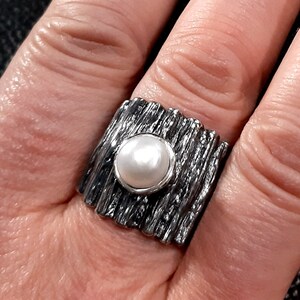 May include: A silver ring with a textured band and a white pearl in the center.
