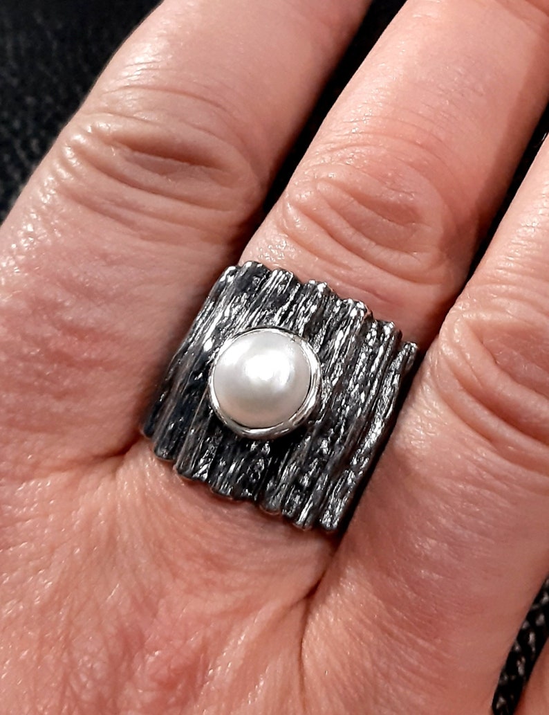 May include: A silver ring with a textured band and a white pearl in the center.