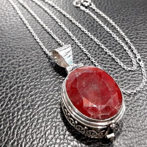Könnte beinhalten: Eine silberne Kettenkette mit einem roten Edelstein-Anhänger. Der Anhänger ist oval geformt und hat einen detaillierten silbernen Rahmen.