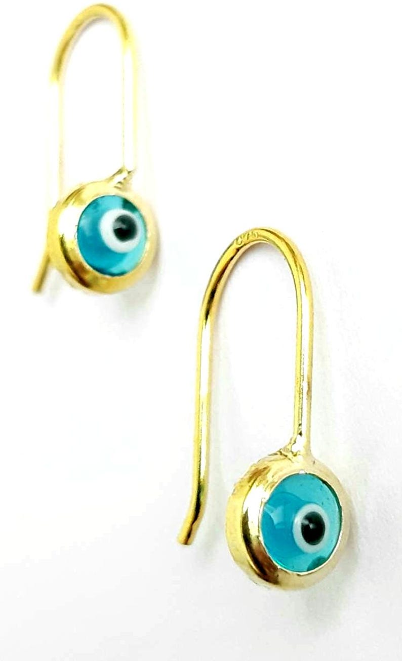 K&ouml;nnte beinhalten: Ein Paar goldene Ohrringe mit blauen b&ouml;sen Augen-Anh&auml;ngern. Die Ohrringe sind aus Gold und haben ein schlichtes Design. Die Anh&auml;nger sind rund und haben eine blaue Emaille-Oberfl&auml;che mit einem wei&szlig;en Auge und einer schwarzen Pupille.