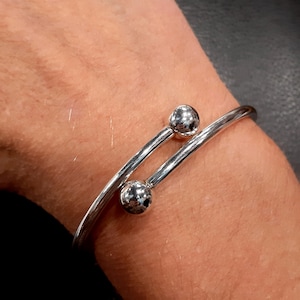 Könnte beinhalten: Silbernes Armreif-Armband mit zwei strukturierten Kugelakzenten. Das Armband ist offen und hat ein schlichtes Design.