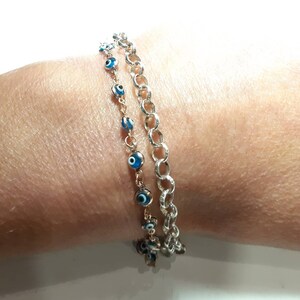 Puede incluir: Dos pulseras en una mu&ntilde;eca. Una pulsera es una cadena de plata con un dise&ntilde;o delicado. La otra pulsera es una cadena de oro con dijes de ojo azul.