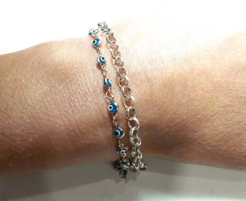 Puede incluir: Dos pulseras en una mu&ntilde;eca. Una pulsera es una cadena de plata con un dise&ntilde;o delicado. La otra pulsera es una cadena de oro con dijes de ojo azul.
