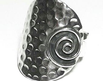 May include: A silver-toned ring with a hammered texture and a spiral design. The ring has a wide, oval face with a textured surface and a raised spiral detail on the side. The metal has a shiny, reflective quality.