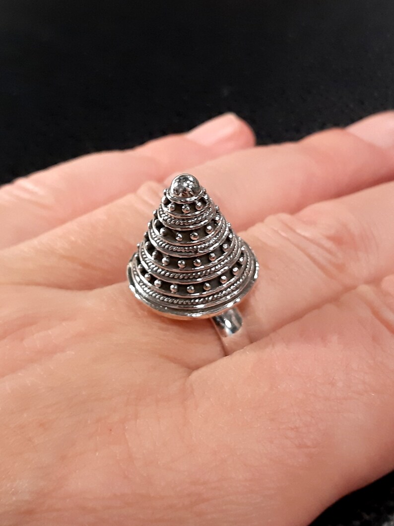 May include: A silver ring with a cone-shaped design featuring multiple layers of intricate detailing. The ring is worn on a finger.