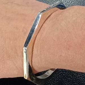 Könnte beinhalten: Ein silberfarbenes, geometrisches Armreif-Armband. Das Armband hat ein flaches, rechteckiges Design mit abgewinkelten Seiten, wodurch eine moderne, minimalistische Ästhetik entsteht. Das Armband wird am Handgelenk getragen.