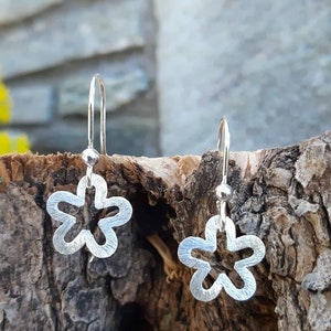 K&ouml;nnte beinhalten: Ein Paar silberfarbene Ohrringe in Blumenform. Jeder Ohrring hat ein f&uuml;nfbl&auml;ttriges Design mit strukturierter Oberfl&auml;che und einfachem Hakenverschluss. Die Ohrringe werden vor einem nat&uuml;rlichen Holzgrund gezeigt.