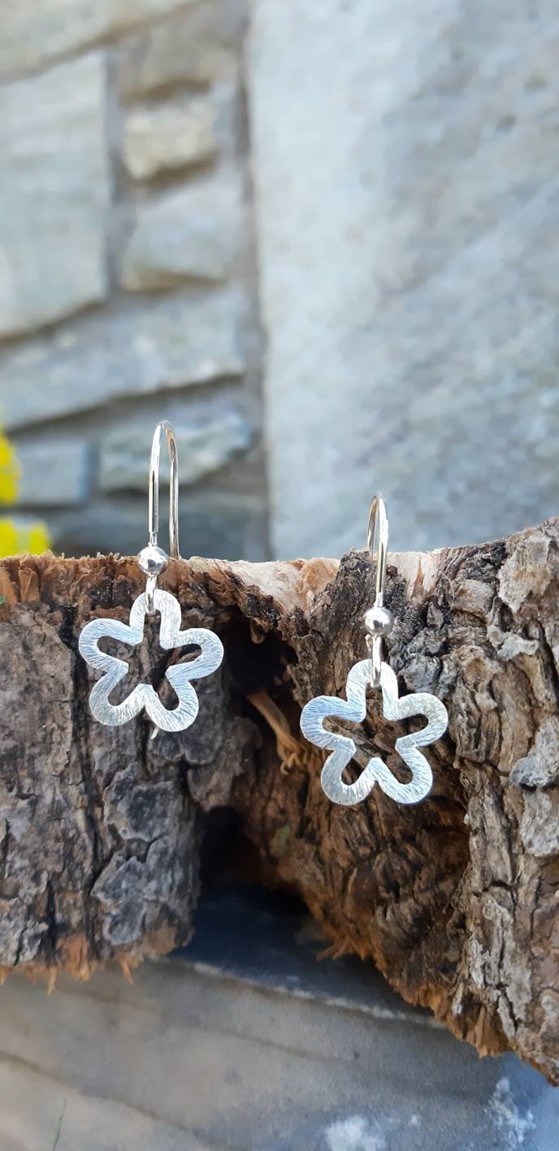 K&ouml;nnte beinhalten: Ein Paar silberfarbene Ohrringe in Blumenform. Jeder Ohrring hat ein f&uuml;nfbl&auml;ttriges Design mit strukturierter Oberfl&auml;che und einfachem Hakenverschluss. Die Ohrringe werden vor einem nat&uuml;rlichen Holzgrund gezeigt.