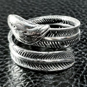 May include: A silver-toned ring featuring a detailed feather design. The ring wraps around the finger with a feather on each end and a stylized bird head at the top. The ring is made of a shiny metal and sits on a dark surface.