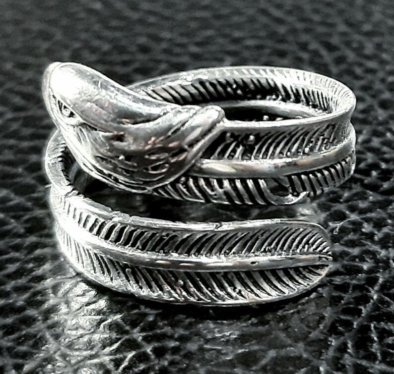 May include: A silver-toned ring featuring a detailed feather design. The ring wraps around the finger with a feather on each end and a stylized bird head at the top. The ring is made of a shiny metal and sits on a dark surface.