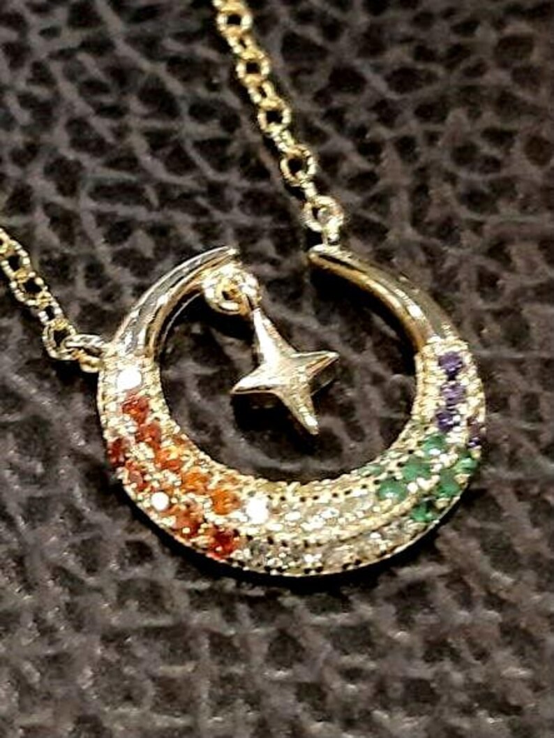May include: A gold crescent moon necklace with a star charm. The crescent moon is adorned with a rainbow of colorful gemstones.