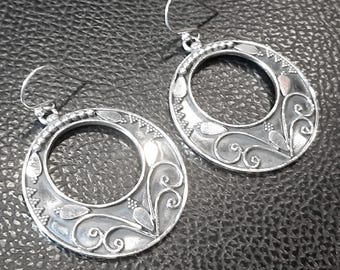May include: A pair of silver-toned hoop earrings. Each earring features an open oval design with intricate floral and leaf patterns. The edges are detailed with small, raised dots and teardrop shapes. The earrings have a hook closure.