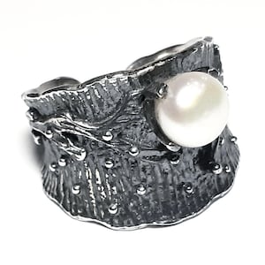 May include: A silver ring with a textured, organic design featuring a large white pearl. The ring has a wide band and a unique, asymmetrical shape.