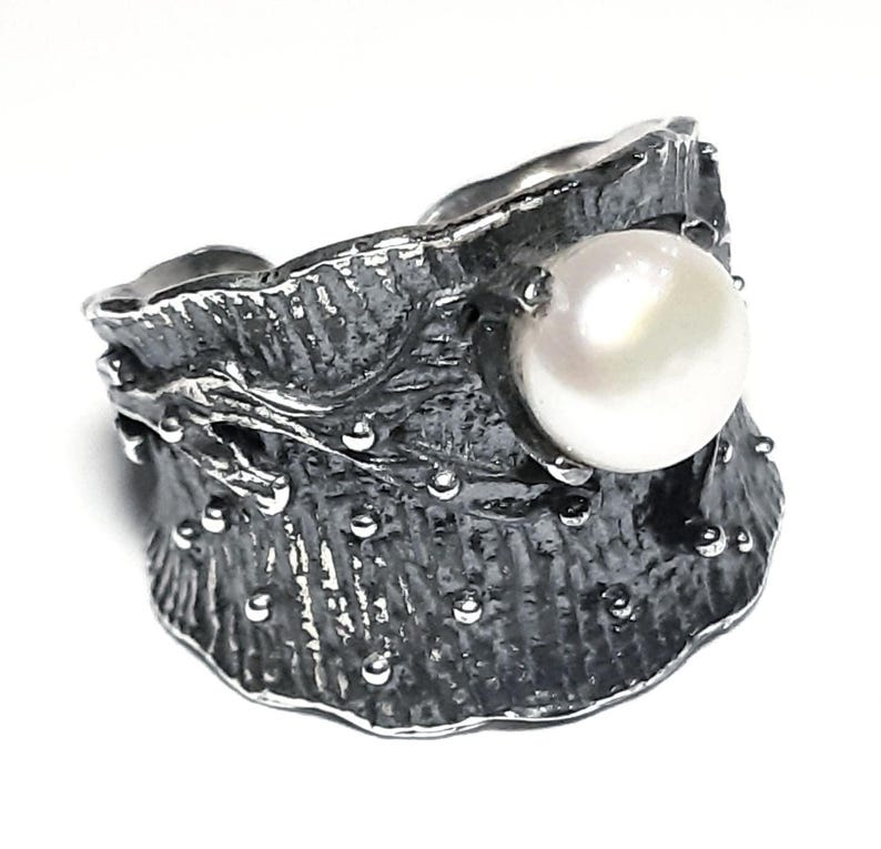 May include: A silver ring with a textured, organic design featuring a large white pearl. The ring has a wide band and a unique, asymmetrical shape.