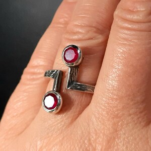 Op de afbeelding: Een zilveren ring met twee rode edelstenen. De ring heeft een geometrisch ontwerp en is aan &eacute;&eacute;n kant open.