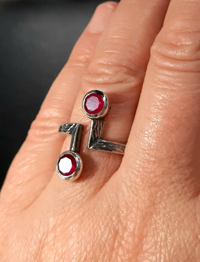 Op de afbeelding: Een zilveren ring met twee rode edelstenen. De ring heeft een geometrisch ontwerp en is aan &eacute;&eacute;n kant open.