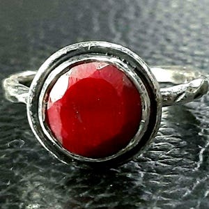 Pu&ograve; includere: Un anello d'argento con una grande pietra preziosa rossa incastonata in una montatura a castone. L'anello ha una fascia testurizzata.