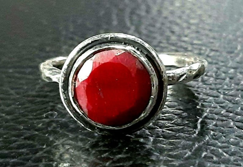 Pu&ograve; includere: Un anello d'argento con una grande pietra preziosa rossa incastonata in una montatura a castone. L'anello ha una fascia testurizzata.