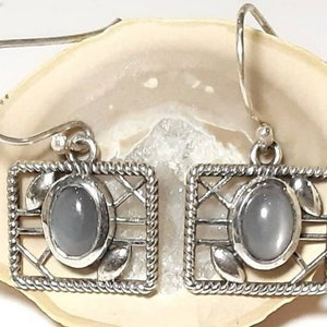 May include: A pair of silver earrings with a gray stone set in a square frame. The frame has a delicate, intricate design.