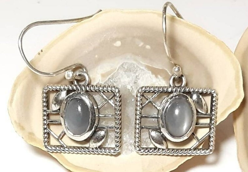 May include: A pair of silver earrings with a gray stone set in a square frame. The frame has a delicate, intricate design.