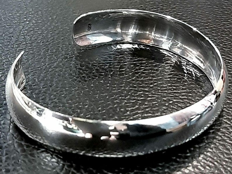 May include: A silver bangle bracelet with a smooth, rounded surface. The bracelet has a hallmark of "925" on the inside.