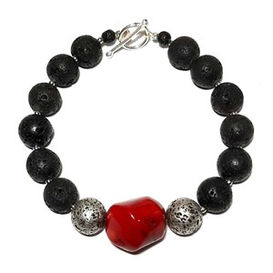 Puede incluir: Un brazalete de cuentas de roca volc&aacute;nica negra con una cuenta de coral rojo y dos cuentas plateadas. El brazalete tiene un cierre de palanca plateado.