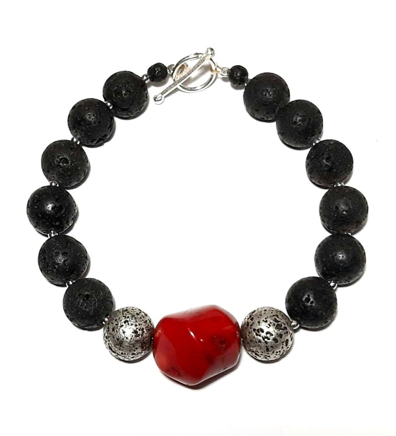 Puede incluir: Un brazalete de cuentas de roca volc&aacute;nica negra con una cuenta de coral rojo y dos cuentas plateadas. El brazalete tiene un cierre de palanca plateado.