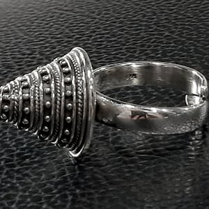 May include: A silver ring with a cone-shaped top. The cone is decorated with a series of concentric circles and dots.
