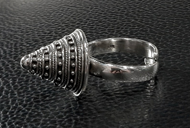 May include: A silver ring with a cone-shaped top. The cone is decorated with a series of concentric circles and dots.