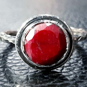 Pu&ograve; includere: Un anello d'argento con una grande pietra preziosa rossa incastonata in una montatura a castone. L'anello ha una fascia testurizzata.
