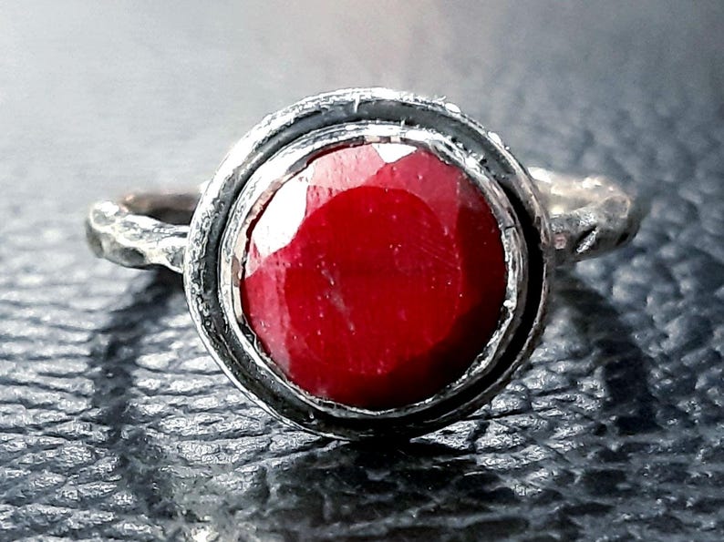 Pu&ograve; includere: Un anello d'argento con una grande pietra preziosa rossa incastonata in una montatura a castone. L'anello ha una fascia testurizzata.