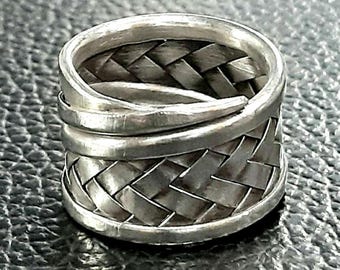 May include: A silver ring with a woven basket design. The ring features a braided pattern around the band and a smooth, wrapped accent. The ring is made of a shiny, metallic material.