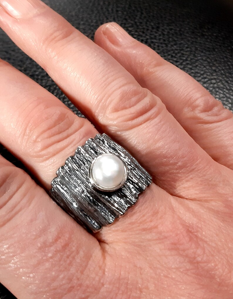 May include: A silver ring with a textured band and a white pearl in the center.