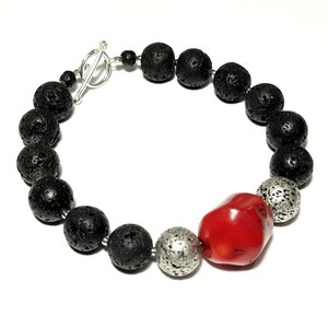 Puede incluir: Un brazalete de piedra de lava negra y cuentas de plata con una gran cuenta de coral rojo. El brazalete tiene un cierre de palanca de plata.