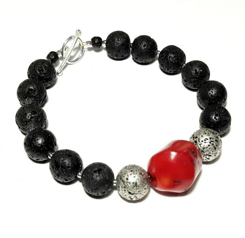 Puede incluir: Un brazalete de piedra de lava negra y cuentas de plata con una gran cuenta de coral rojo. El brazalete tiene un cierre de palanca de plata.