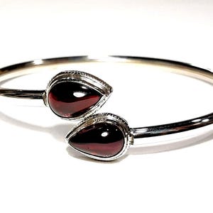 Könnte beinhalten: Ein silbernes Armreif-Armband mit zwei tropfenförmigen roten Edelsteinen.
