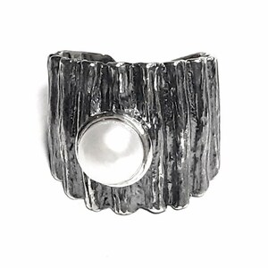 May include: A silver ring with a textured, wood-like design. The ring has a circular cutout that features a white pearl.