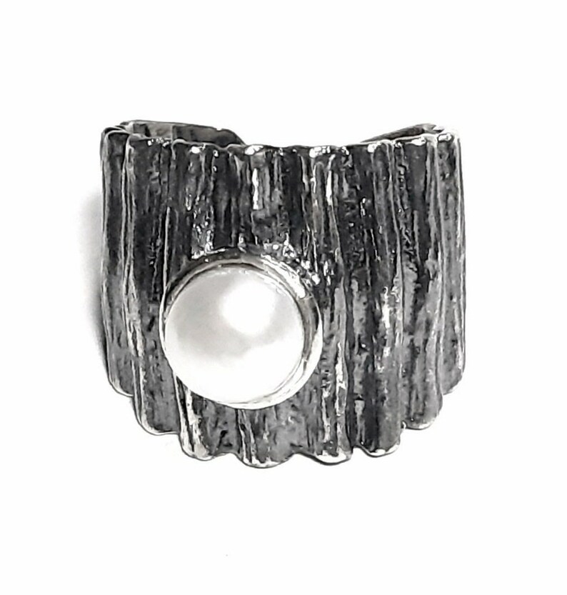 May include: A silver ring with a textured, wood-like design. The ring has a circular cutout that features a white pearl.