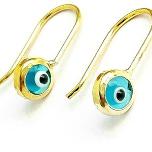 K&ouml;nnte beinhalten: Ein Paar goldene Ohrringe mit einem blauen b&ouml;sen Blick-Anh&auml;nger. Die Ohrringe sind aus Gold gefertigt und haben ein schlichtes Design. Der b&ouml;se Blick-Anh&auml;nger ist ein beliebtes Symbol f&uuml;r Schutz und Gl&uuml;ck.