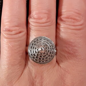 May include: A silver ring with a circular design featuring concentric circles and a small, raised center. The ring is worn on a finger.