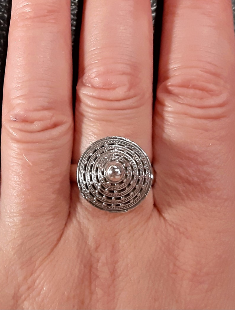 May include: A silver ring with a circular design featuring concentric circles and a small, raised center. The ring is worn on a finger.