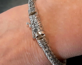 May include: A silver bracelet featuring a woven chain link design and a cylindrical clasp mechanism. The clasp is decorated with intricate embossed patterns and includes a small lever for securing the closure on the wrist.
