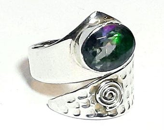 May include: A silver-toned bypass ring featuring a polished oval-shaped stone with iridescent green and purple highlights. The band has a hammered texture and a prominent spiral design on one side, creating a wrap-around style with an open front.