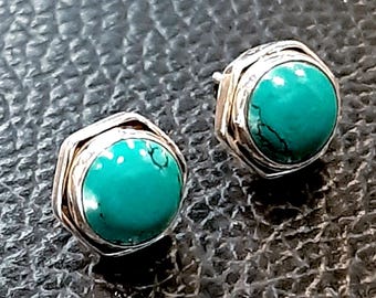 May include: A pair of turquoise stud earrings. Each earring features a round, vibrant turquoise stone set in a silver-colored, square-shaped bezel. The turquoise stones have dark veining. The earrings are displayed on a textured, dark surface.