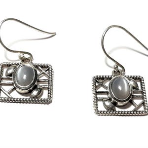 May include: Silver earrings with a white oval gemstone set in a square frame with a delicate design.