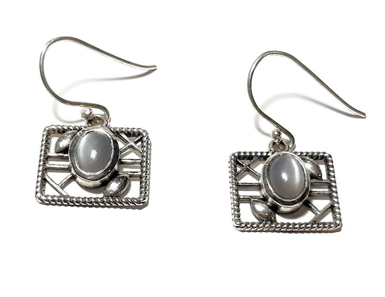 May include: Silver earrings with a white oval gemstone set in a square frame with a delicate design.